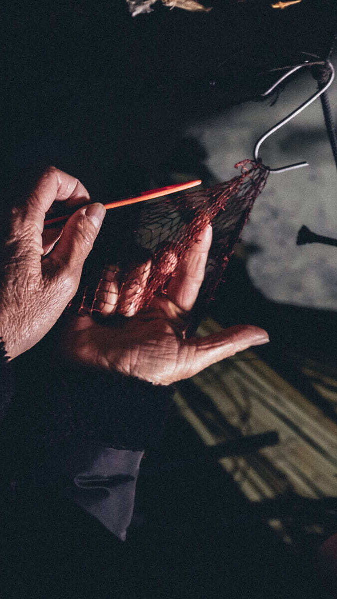 Primer plano de las manos de una mujer reparando una red de pesca con una aguja o herramienta de tejer, bajo una luz tenue.